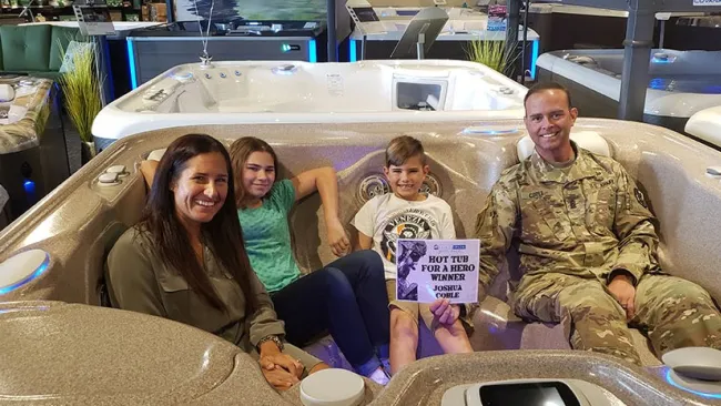 Family enjoying a hot tub demonstration in a showroom with a military father holding a sign.
