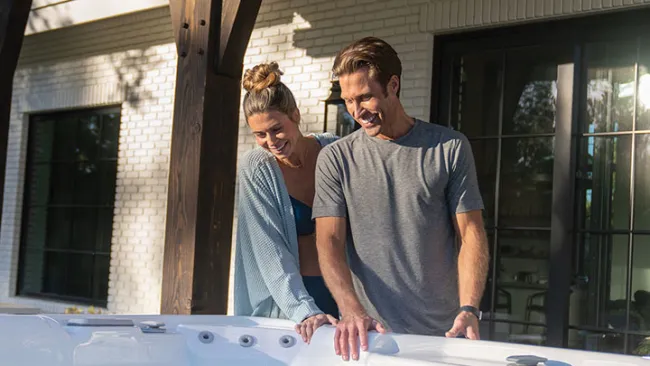 Smiling couple checking water in an outdoor white hot tub on a sunny day near a modern home.