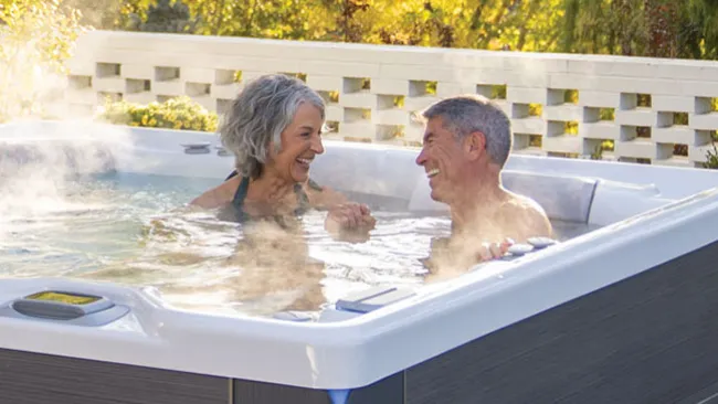 Smiling mature couple enjoying a steaming hot tub outdoors on a sunny day surrounded by greenery.