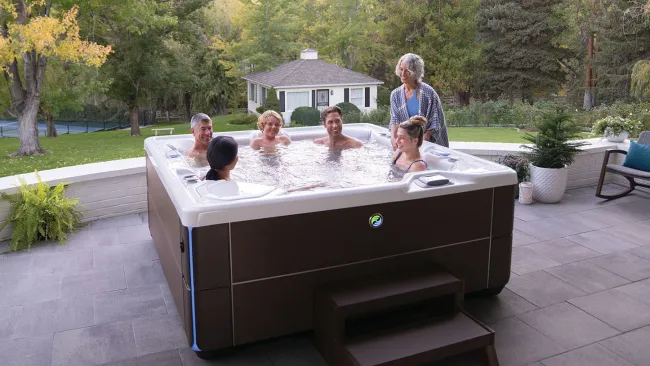Six adults enjoying a large outdoor hot tub on a patio surrounded by trees and a garden house.