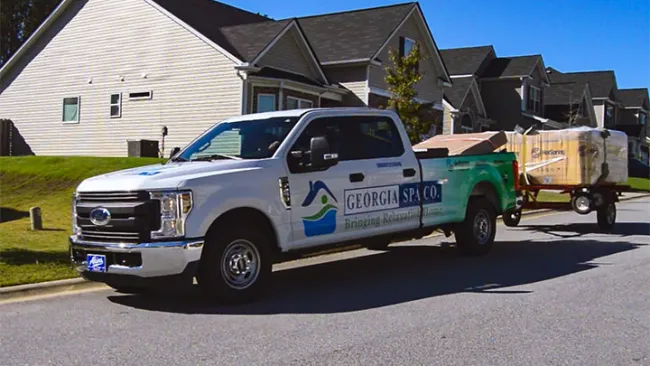 White Georgia Spa Co. pickup truck towing a trailer loaded with spa equipment parked in a suburban neighborhood.