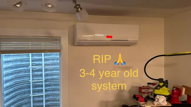 Indoor wall-mounted air conditioning unit above a desk with tools and text saying RIP 3-4 year old system.