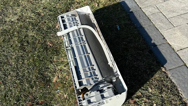 Old broken air conditioner unit discarded on grass next to paved sidewalk in daylight.