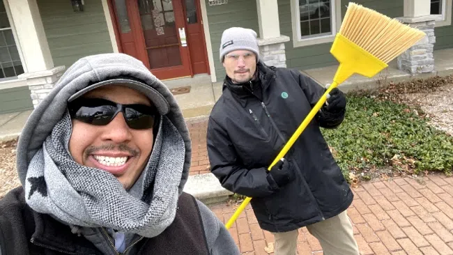 Two men dressed in winter clothing smiling outside a house with one holding a large yellow broom.