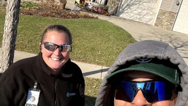 Two smiling people wearing sunglasses, outdoors near a residential house, one giving a thumbs up in bright sunlight.