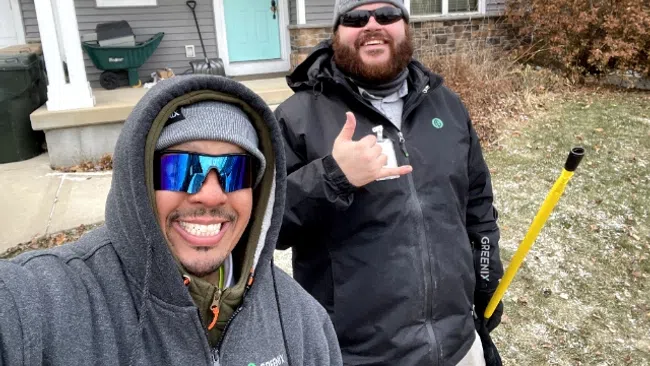 Two men in winter clothing smiling outdoors in front of a house with a broom on a cold day