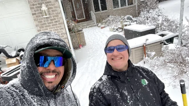 Two people smiling in winter jackets and sunglasses while standing outside in a snowy residential yard during snowfall
