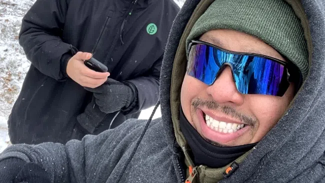 Two men dressed in winter jackets and hats smile outdoors in a snowy yard with houses in the background.