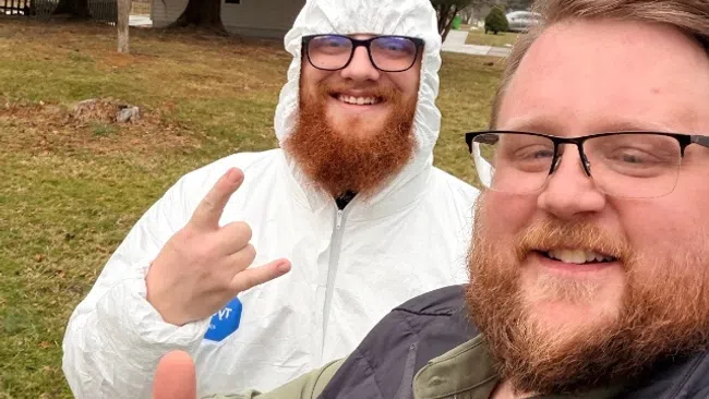 Two men smiling outdoors, one in a white protective suit showing rock sign, the other giving a thumbs up.