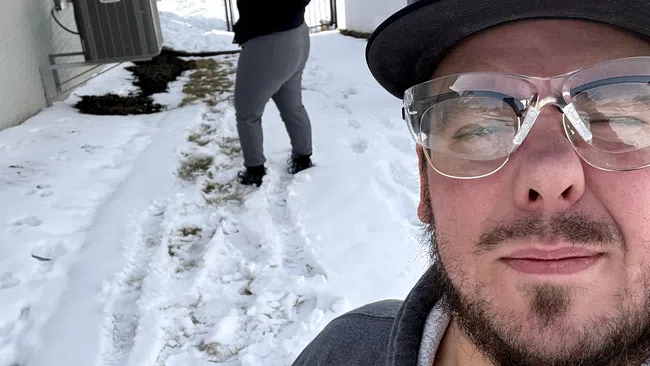 Man in glasses and hoodie takes selfie outside in snow with woman in background holding long broom or pole.