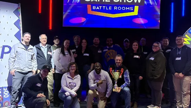 Group of people posing with a trophy in front of a Game Show Battle Rooms screen with colorful lights.