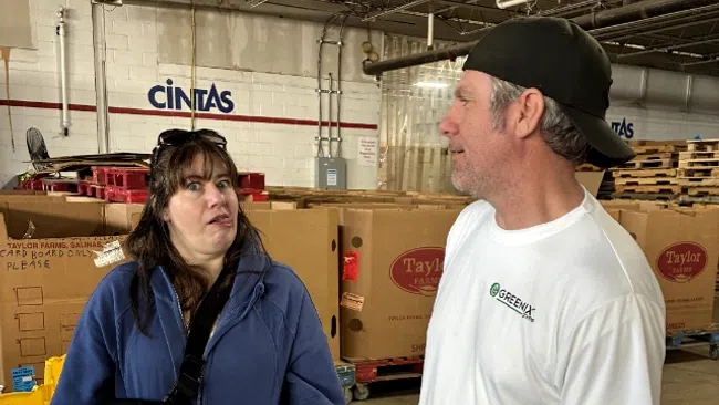 Two people talking inside a warehouse with stacked cardboard boxes and industrial shelving in the background