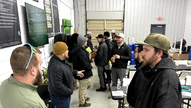 Group of men in casual outdoor clothing standing and talking inside a warehouse or meeting space.