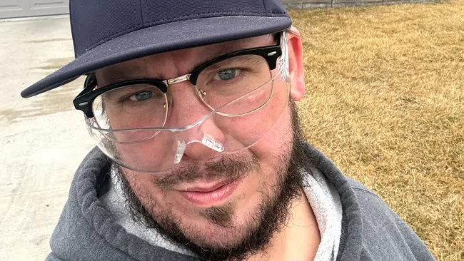 Man wearing safety glasses and a cap takes a selfie outside a house with a worker using a leaf blower in the background.
