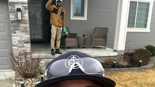 Man wearing glasses and a cap takes a selfie in front of a house with another man waving from the porch.
