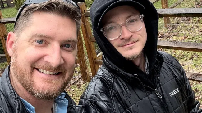 Two men in black jackets, one wearing a hood, smiling outdoors near a wooden fence and deck on a rainy day.