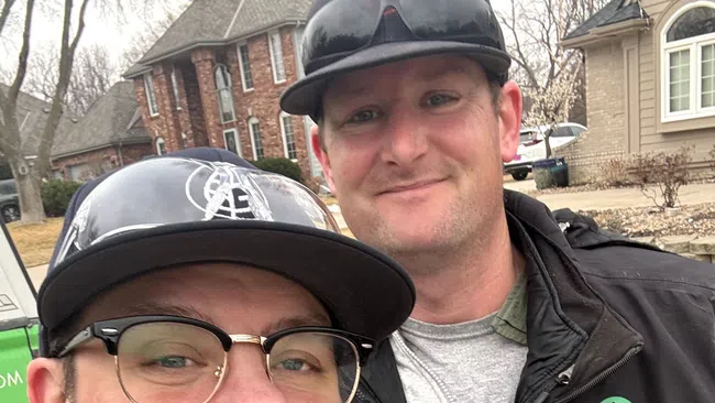 Two men smiling outdoors wearing casual jackets and caps with residential houses in the background on a cloudy day.