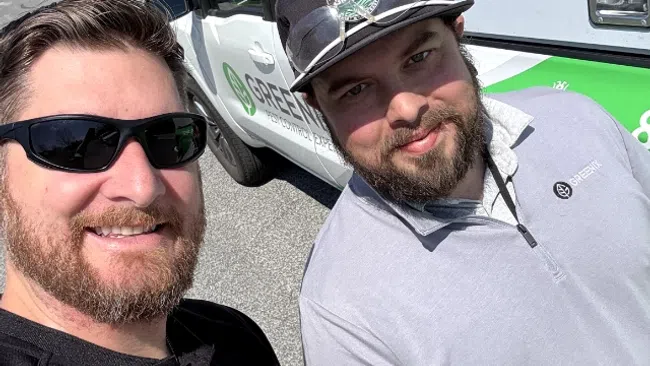 Two men smiling and posing for a selfie next to a pest control van in a sunny suburban street.