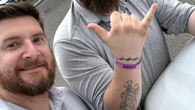 Two men with beards taking a selfie outdoors, one wearing reflective sunglasses and making a hand gesture, the other smiling.