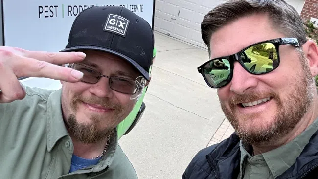 Two pest control workers in Greenix uniforms pose smiling outdoors in front of a service van and garage door.