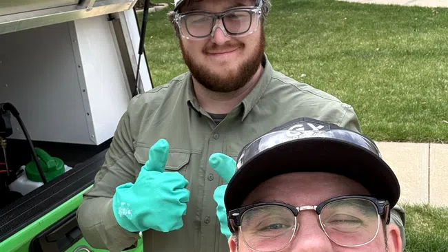 Two men smiling near a green Phoenix Pest Control truck, one wearing green gloves and giving thumbs up, outdoors in a residential area.