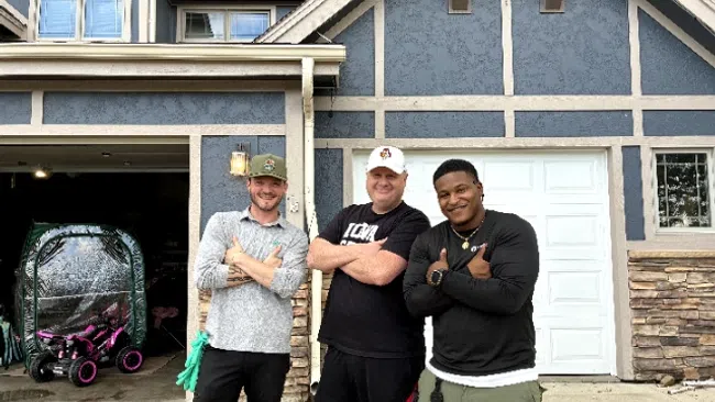 Three men smiling and posing with crossed arms outside a blue house with garage doors and stone accents.