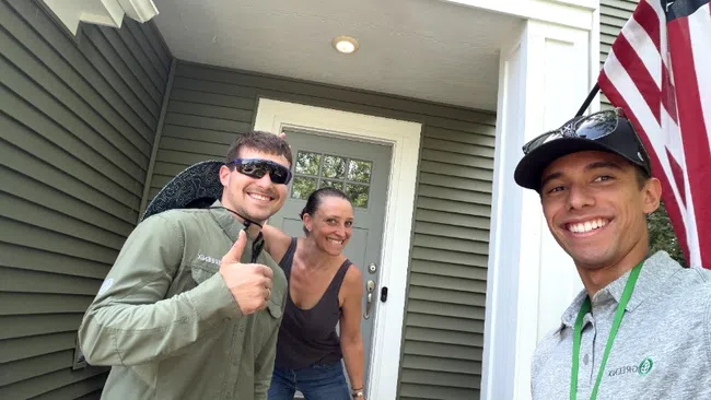 Three smiling people posing outside a house entrance with an American flag and giving a thumbs up on a sunny day.