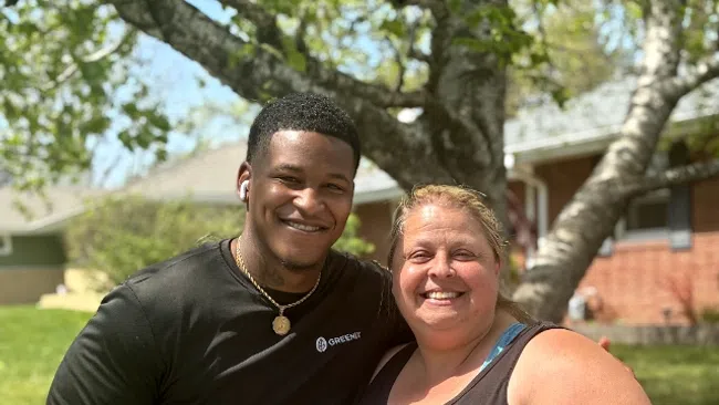 Smiling man and woman posing outdoors in casual sportswear with a tree and houses in the background.
