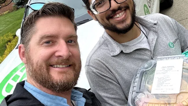 Two smiling men posing in front of a green and white service truck, one holding a food container and making a hand gesture.