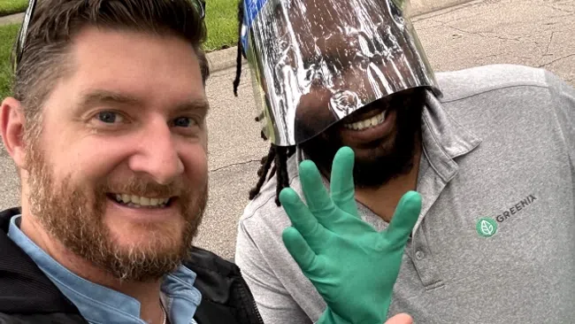 Two men smiling outdoors, one wearing a face shield and green gloves, the other making a hand gesture in casual attire.