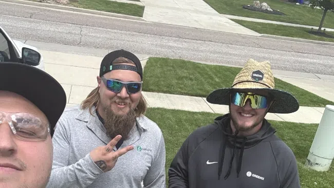Three landscapers wearing work gear pose for a selfie on a suburban sidewalk with green lawns and houses in the background.