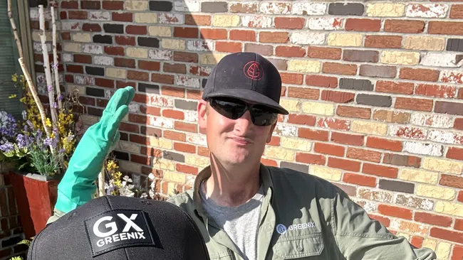 Two men wearing GX Greenix hats and safety glasses posing in front of a brick wall with plants on a sunny day.