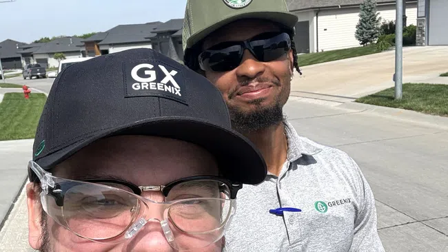 Two men wearing Greenix branded hats and shirts smiling outdoors in a suburban neighborhood on a sunny day.