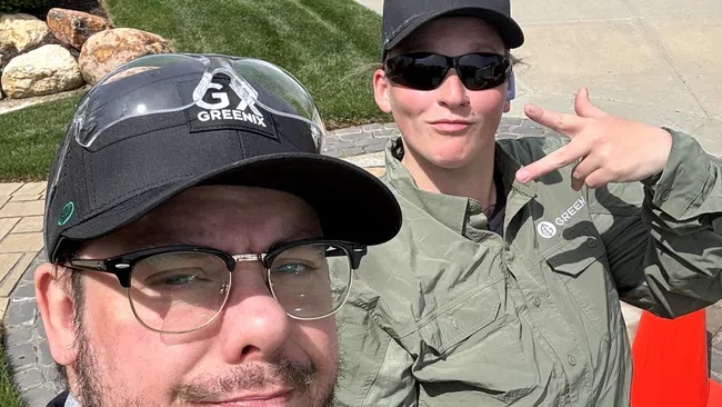 Two Greenix employees wearing uniforms and caps posing outdoors with snacks and drinks on a sunny day.