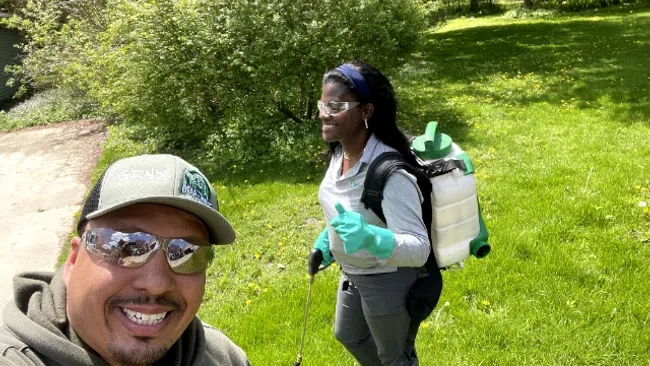 Two outdoor workers in protective gear with backpack sprayers on a green lawn on a sunny day