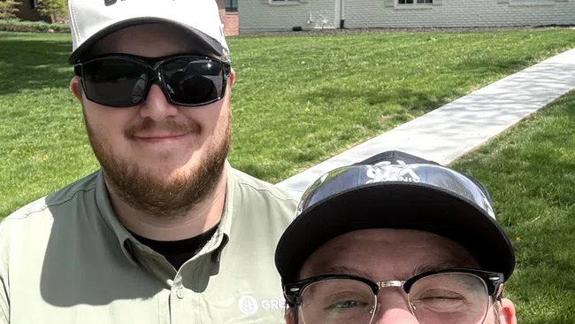 Two men outdoors smiling, one wearing a GRNX cap and sunglasses, the other wearing glasses and a cap, with a house in background.