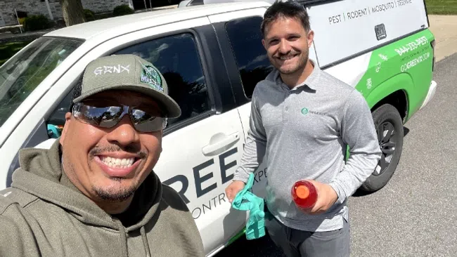 Two pest control technicians smiling beside a branded white and green service truck on a residential street.