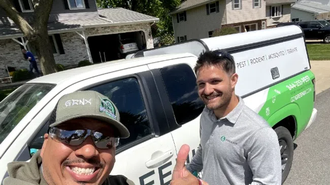 Two pest control workers smiling in front of a white and green service truck on a sunny residential street.