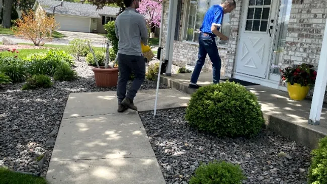 Two men approaching a suburban house entrance on a sunny day with landscaped garden and stone path.