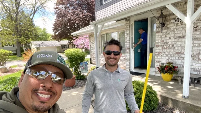 Two men smiling outdoors in front of a suburban house, one holding a yellow pole, another painting a door inside.