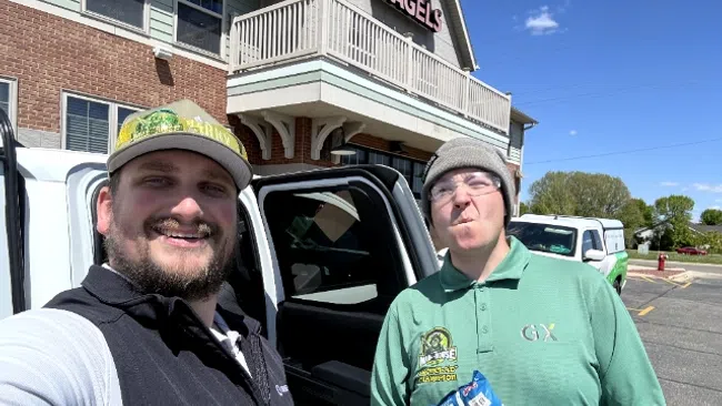 Two men smiling outdoors by a white pickup truck near a brick building with a sign that reads Bagels