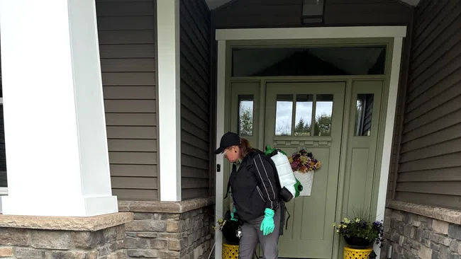 Person wearing gloves and backpack sprayer applying treatment on a house front porch with stone walls.