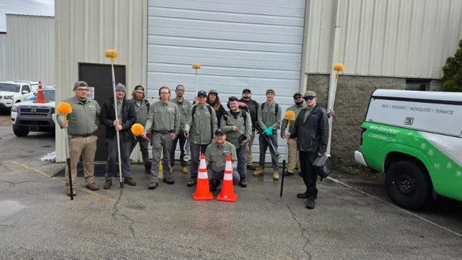 Group of pest control workers in uniform standing outside a building with equipment and company vehicle.