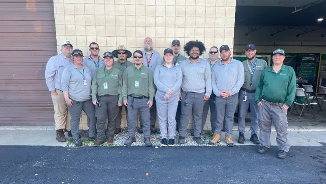Group of fifteen outdoor workers and staff standing in front of a beige building wall in work uniforms.