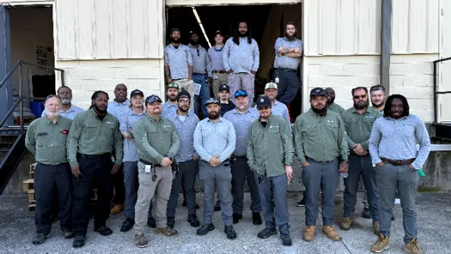 Group of diverse men in work uniforms posing outside beige industrial building on concrete ground.