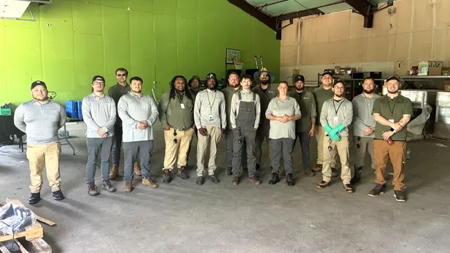 Group photo of fifteen warehouse workers standing inside a large industrial warehouse with green walls.