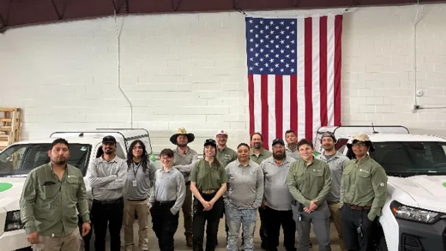 Group of diverse workers standing in front of two white trucks with a large American flag on the wall behind.