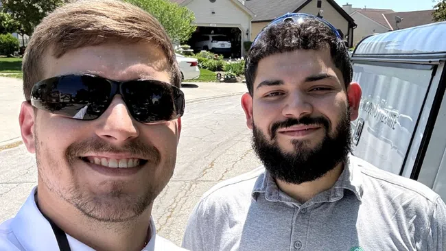 Two men wearing Greenix uniforms smile outdoors on a sunny day in a suburban neighborhood.