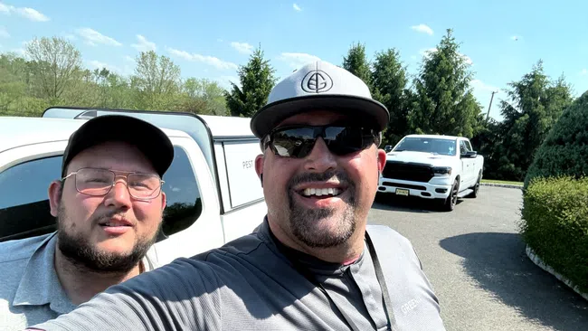 Two men smiling outdoors near white vehicles on a sunny day with trees and clear blue sky