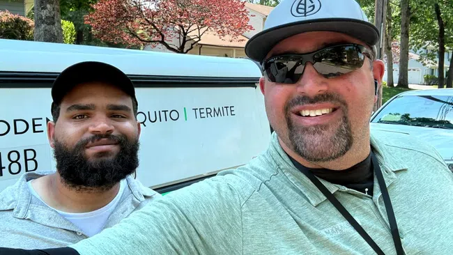 Two pest control workers smiling outdoors with van showing mosquito and termite services in background.